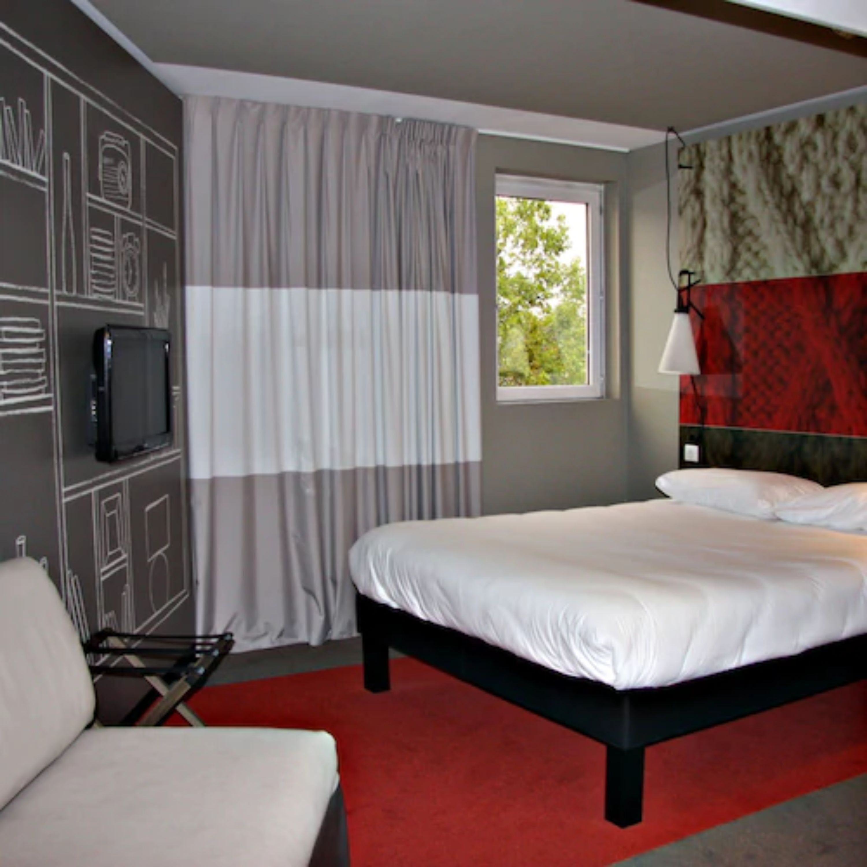 A hotel room with a bed, a TV, a window with curtains, and a red carpet.