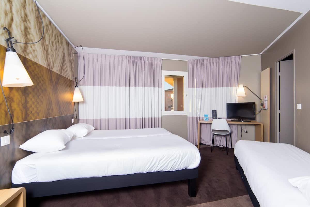 Une chambre d’hôtel avec deux lits, un bureau avec une télévision et une fenêtre avec des rideaux.
