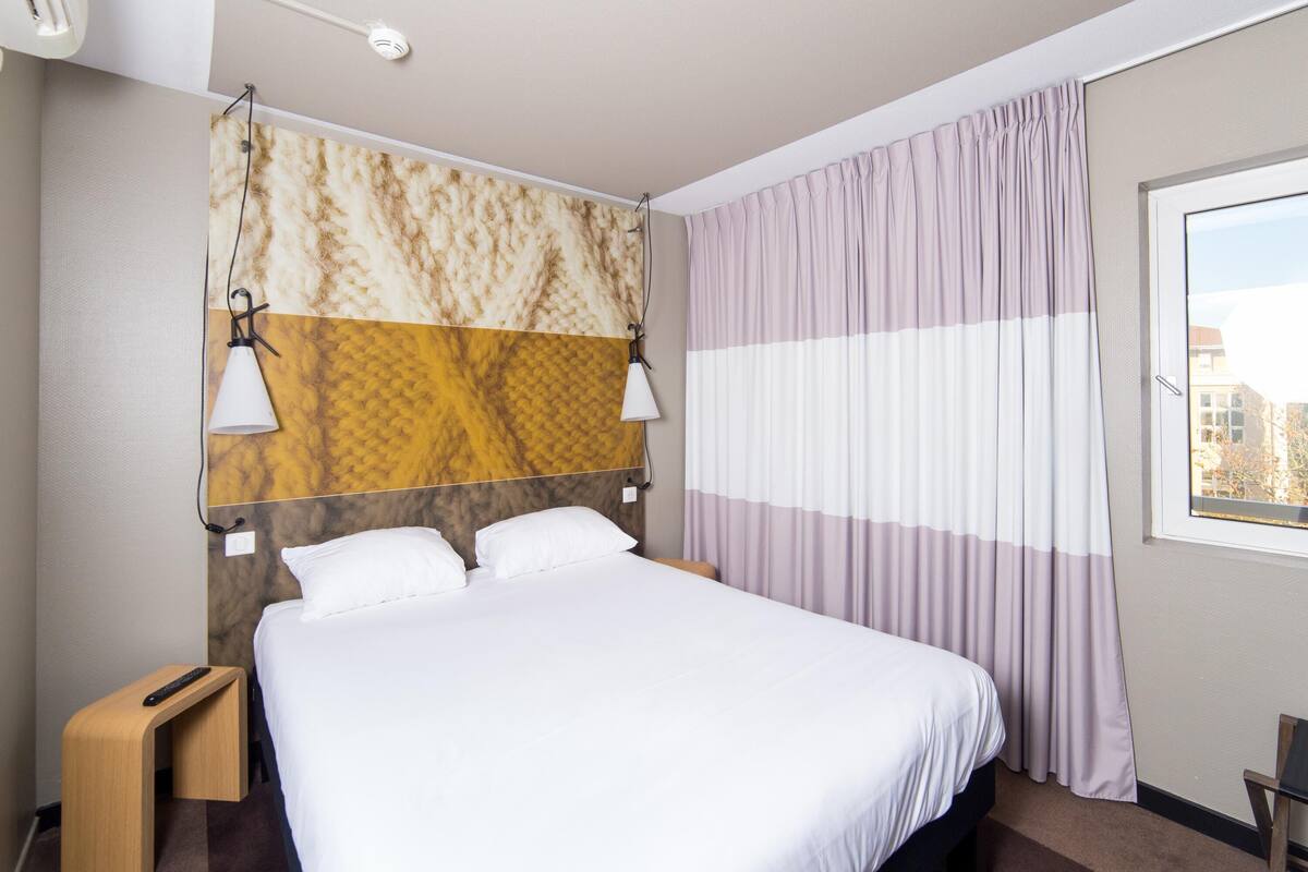 Une chambre d’hôtel comprenant un lit, deux lampes de chevet, une tête de lit au motif texturé, une fenêtre avec des rideaux et une petite table de chevet en bois.