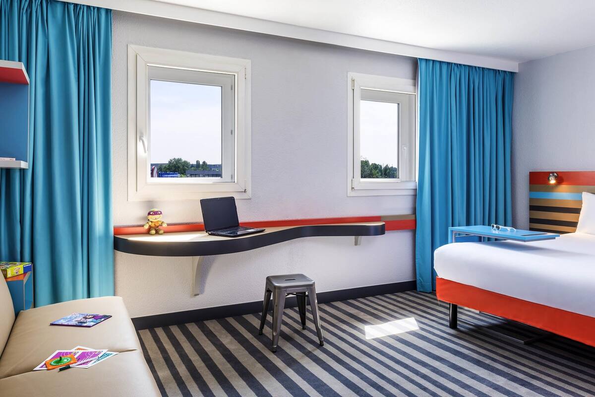 Ett hotellrum med en säng, ett skrivbord med en bärbar dator, en stol, en liten pall och ett fönster med gardiner.