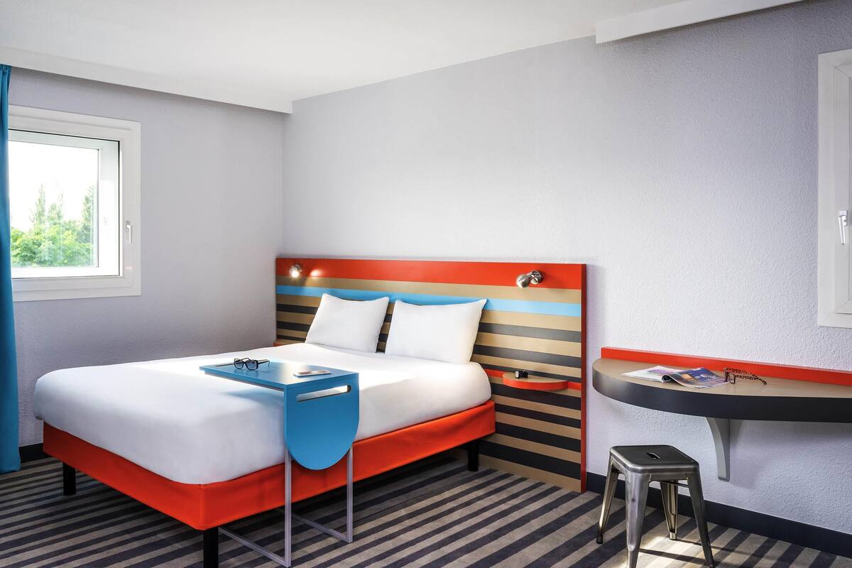 A hotel room with a bed, a desk, a chair, a window, and a wall-mounted shelf.