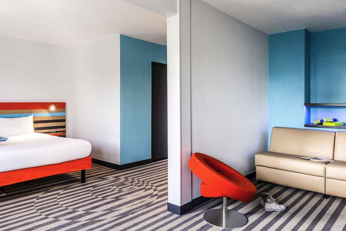 Quarto de hotel com cama, uma cadeira vermelha, sofá e uma pequena cozinha compacta.