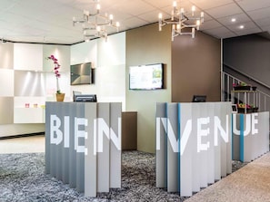 A reception area with 'BIENVENUE' signage, a flat-screen TV, a potted orchid, and a staircase in the background.