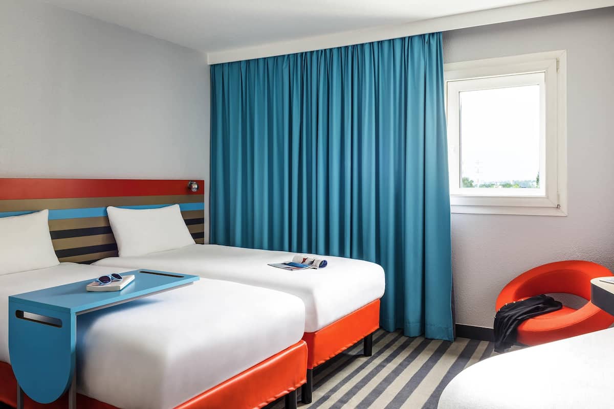 Quarto de hotel com duas camas, uma janela com cortinas, uma mesinha e uma cadeira.