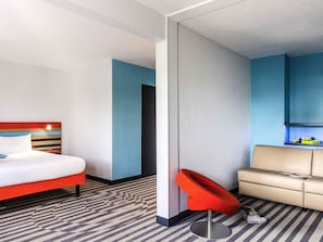 A hotel room with a bed, a red chair, a sofa, and a small kitchenette. - ibis Styles Antony Paris Sud (Antony)