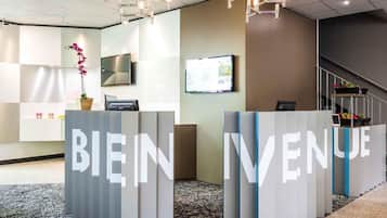 A reception area with 'BIENVENUE' signage, a flat-screen TV, a potted orchid, and a staircase in the background.
