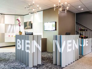 Una zona de recepción con letrero que dice 'BIENVENUE', un televisor de pantalla plana, una orquídea en maceta y una escalera al fondo.
