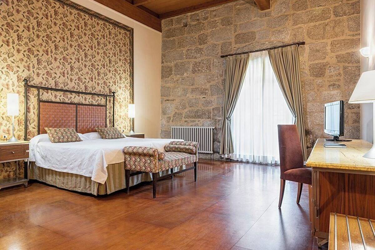 A bedroom with a large bed, a desk, and a chair. The room has stone walls and a wooden floor.
