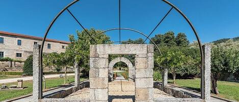 A stone archway structure with a metal framework, surrounded by a paved area and greenery.
