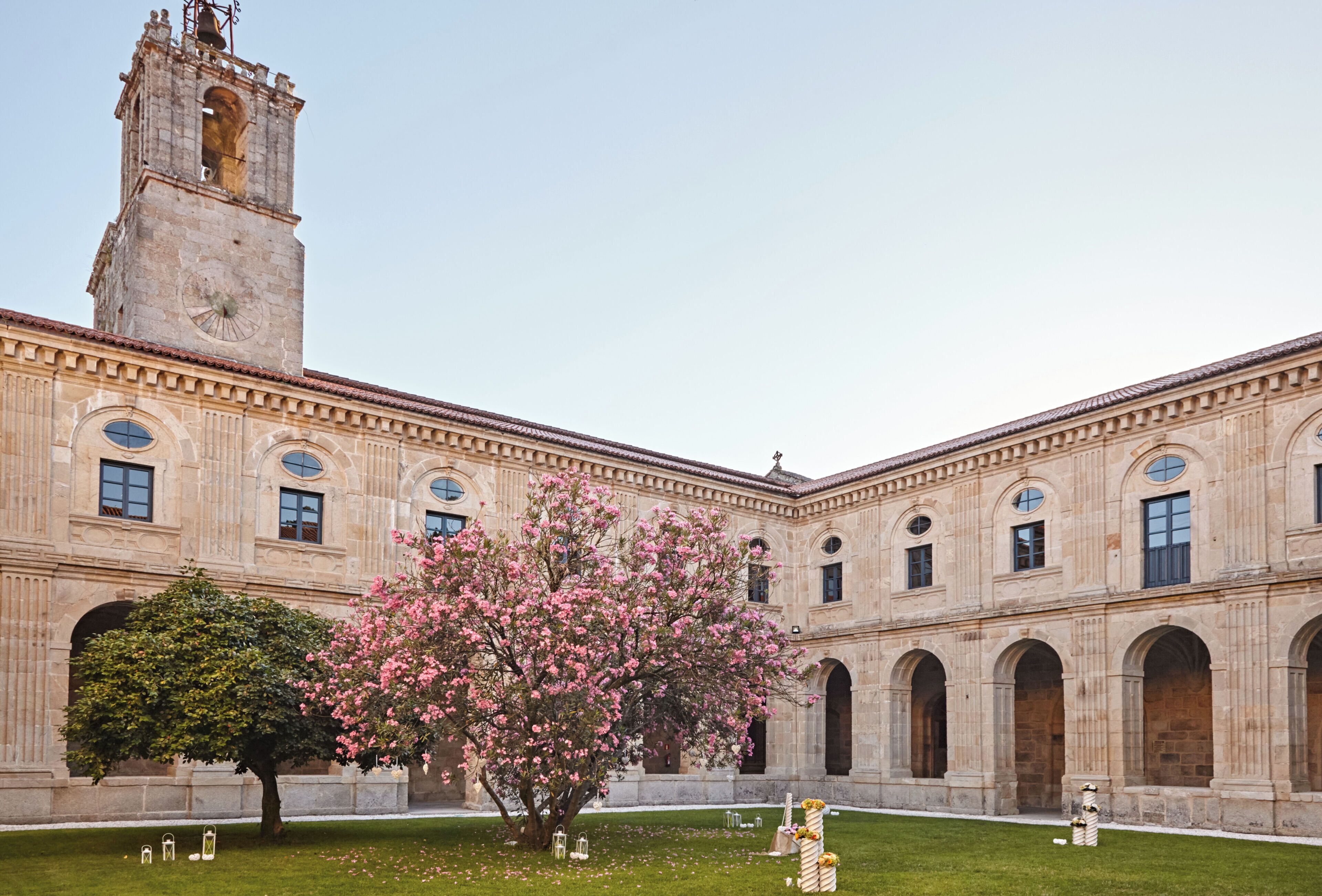 Un edificio histórico con una torre de reloj, ventanas de arco y un patio con un árbol y flores.