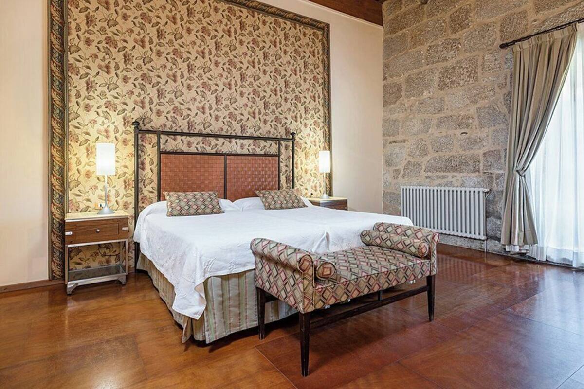 A bedroom with a large bed, two bedside tables, a bench, a stone wall, and a window with curtains.