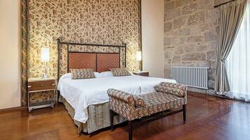 Un dormitorio con una cama grande, dos mesitas de noche, un banco, una pared de piedra y una ventana con cortinas.