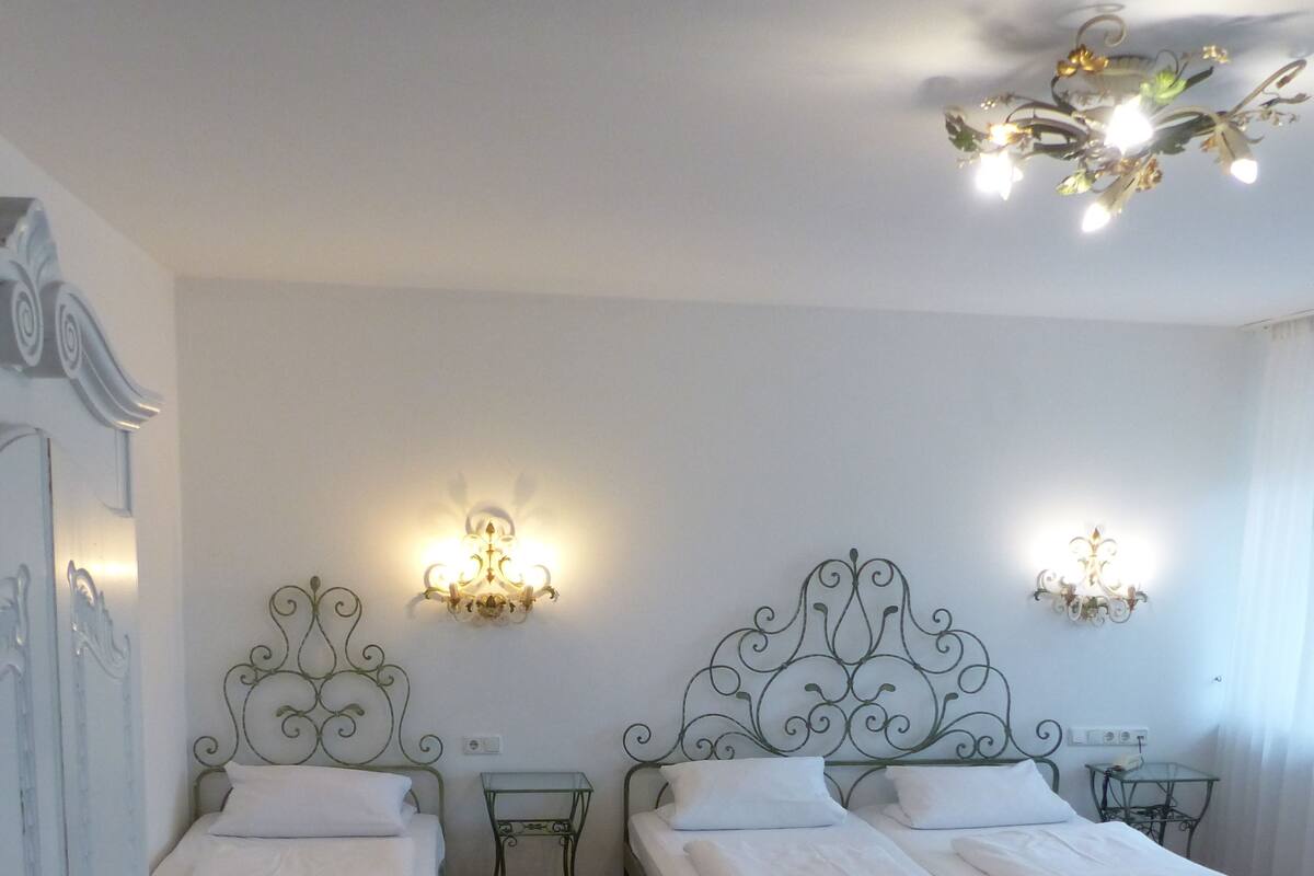 A double room with two beds, each with a metal headboard and white bedding. There are two wall-mounted light fixtures and a ceiling light fixture.