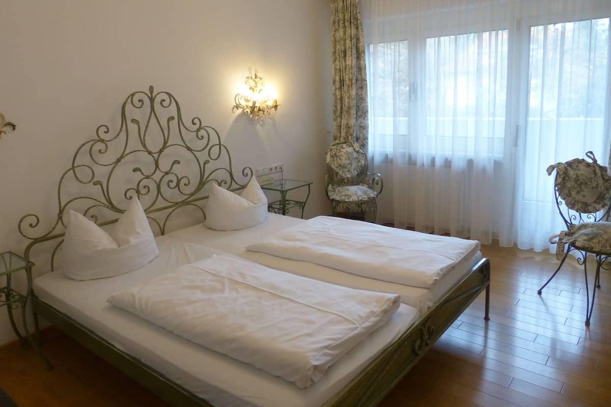 A double bed with white linens, a decorative metal headboard, a wall-mounted light fixture, a chair with a floral pattern, and a window with sheer curtains.