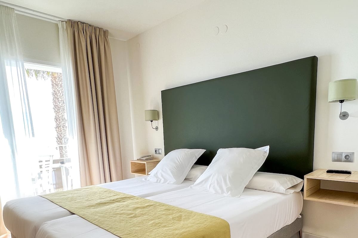 A hotel room with a large bed, wooden flooring, a green headboard, and a window with curtains.