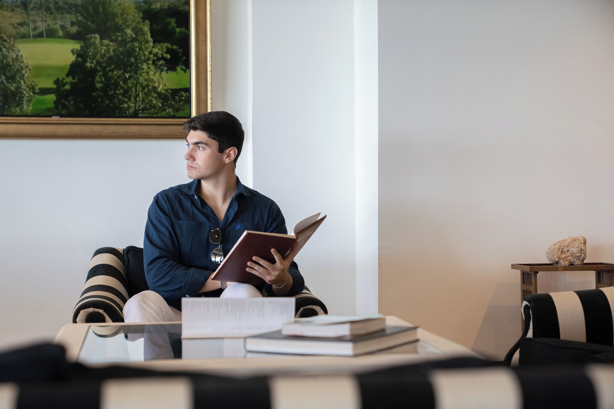 Eine Person sitzt auf einem gestreiften Sofa in einem Raum, an der Wand hängt ein Gemälde eines Golfplatzes, ein kleiner Holztisch mit einem Stein und mehrere Bücher auf einem Couchtisch.