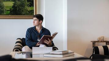 Eine Person sitzt auf einem gestreiften Sofa in einem Raum, an der Wand hängt ein Gemälde eines Golfplatzes, ein kleiner Holztisch mit einem Stein und mehrere Bücher auf einem Couchtisch.