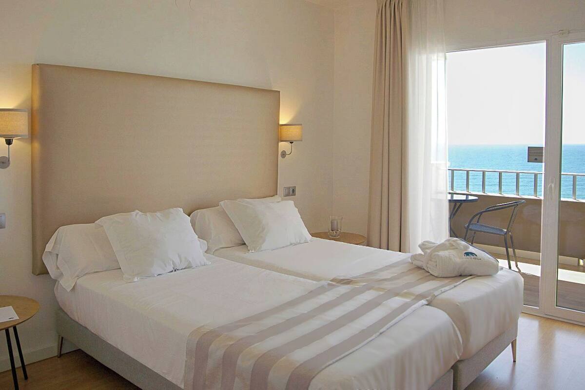 A hotel room with a large bed, two bedside tables, a chair, and a balcony with a view of the sea.