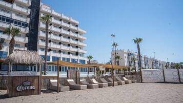 Un hotel frente a la playa con varios pisos, terraza al aire libre y palmeras.