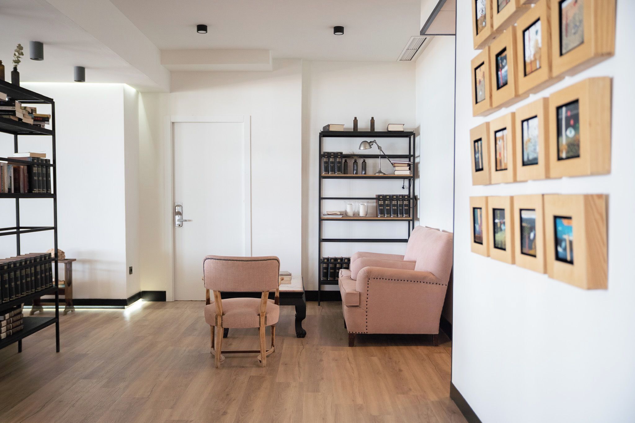 Una sala moderna con piso de madera, dos sillones rosados, una estantería y una pared con cuadros enmarcados.