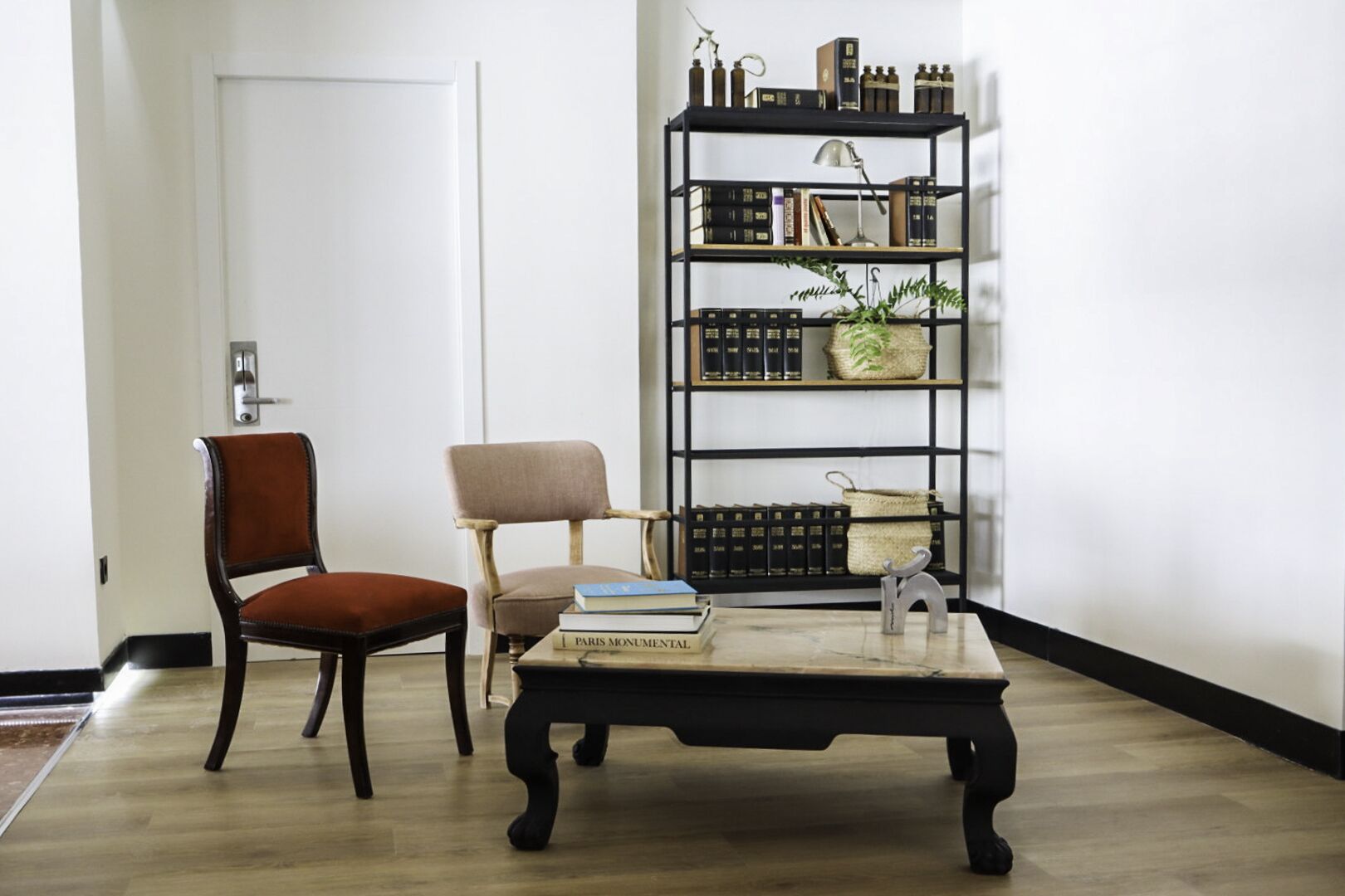 Ein Zimmer mit Holzboden, einem braunen Stuhl, einem beigen Sessel, einem schwarzen Couchtisch mit Büchern und einem schwarzen Bücherregal mit Büchern und Dekoration.