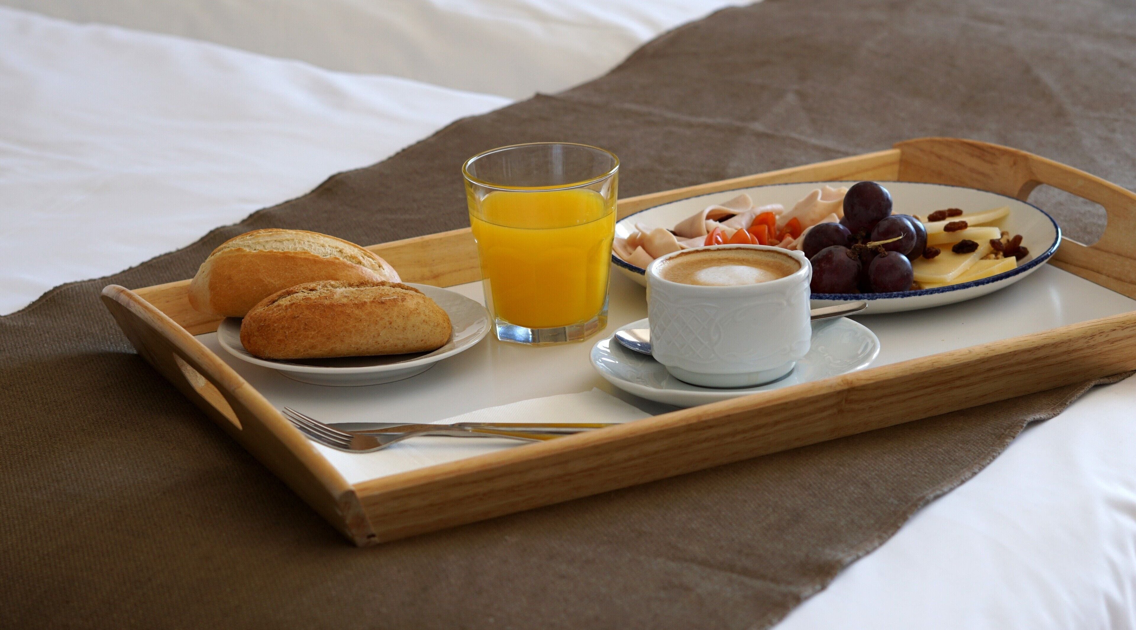 Bandeja de desayuno con pan, zumo de naranja, café y una fuente de fruta y queso sobre una cama.