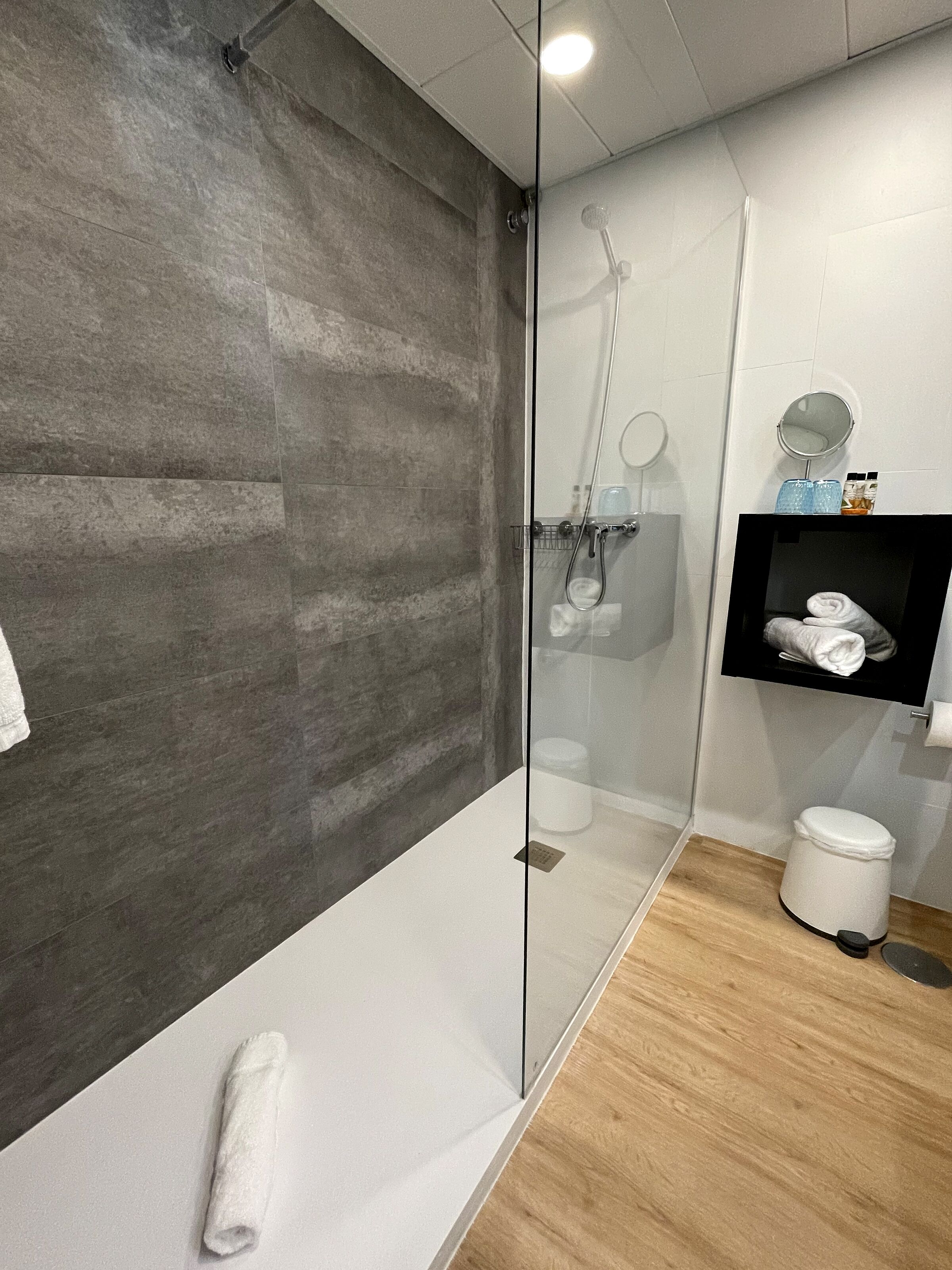 Un baño moderno con ducha de vidrio, piso de madera y tocador blanco con espejo y perchero.