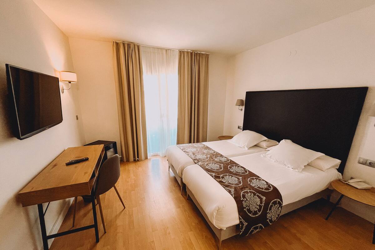 A hotel room with a bed, a desk, a chair, a TV, and a window with curtains.