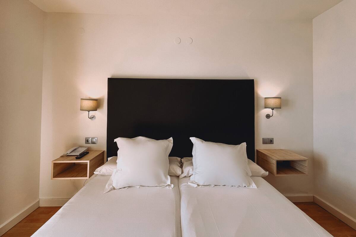 A double bed with white linens, two bedside tables, a black headboard, and wall-mounted lamps.