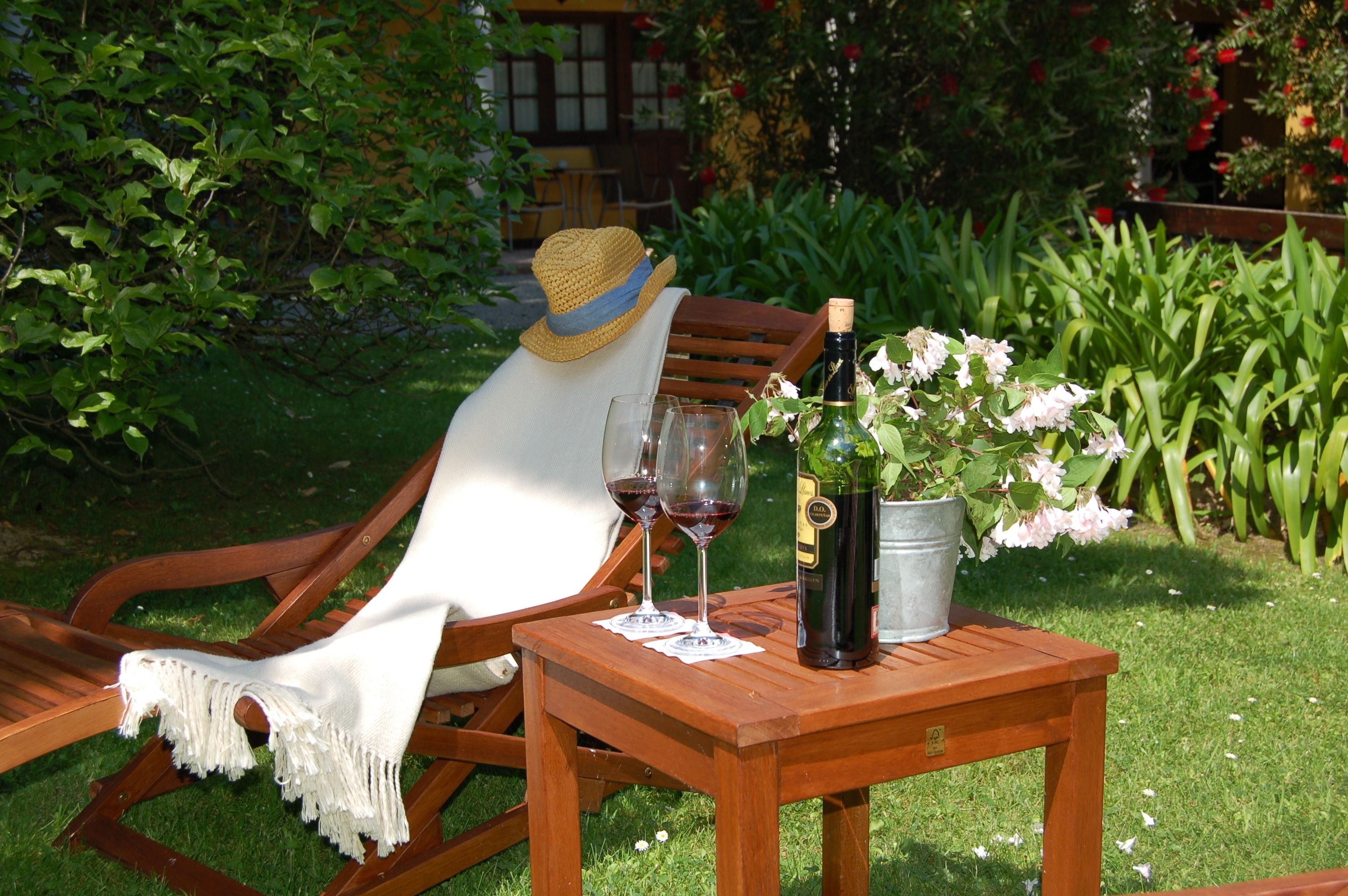 Ein Holzsessel mit weißem Stoffbezug und ein Strohhut, ein Holztisch mit einer Weinflasche und Gläsern sowie ein Blumentopf mit Blumen.
