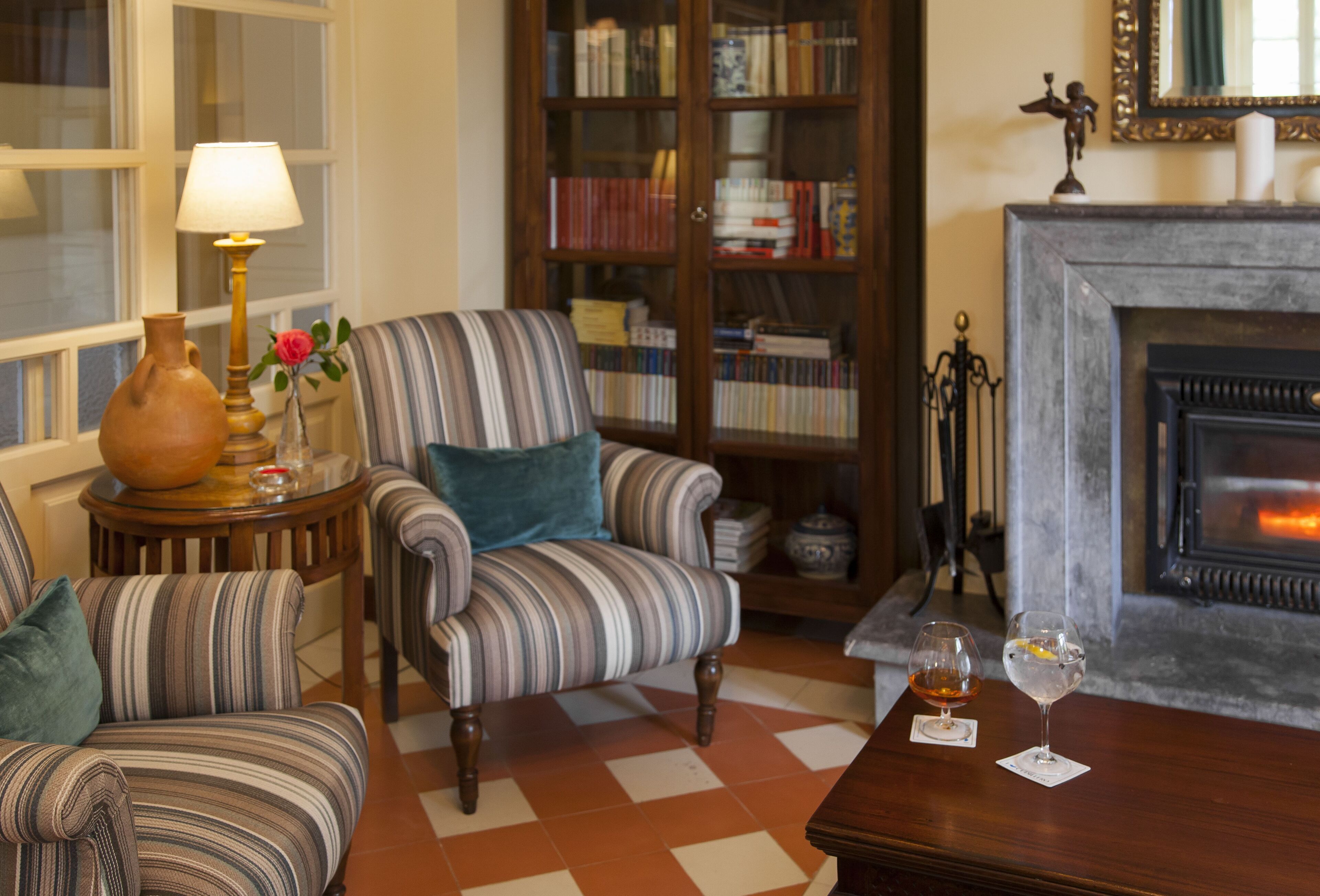 A cozy living room with striped armchairs, a fireplace, a bookshelf, and a table with glasses.