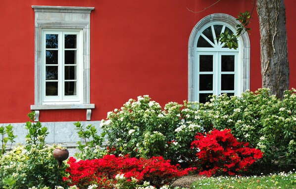 A red building with two white-framed windows and a variety of flowering plants in the foreground. - Casona de la Paca (Cudillero)