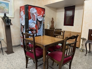 Uma área de refeições com uma mesa de madeira e cadeiras com almofadas vermelhas, uma geladeira Coca-Cola e uma estátua em um pedestal.