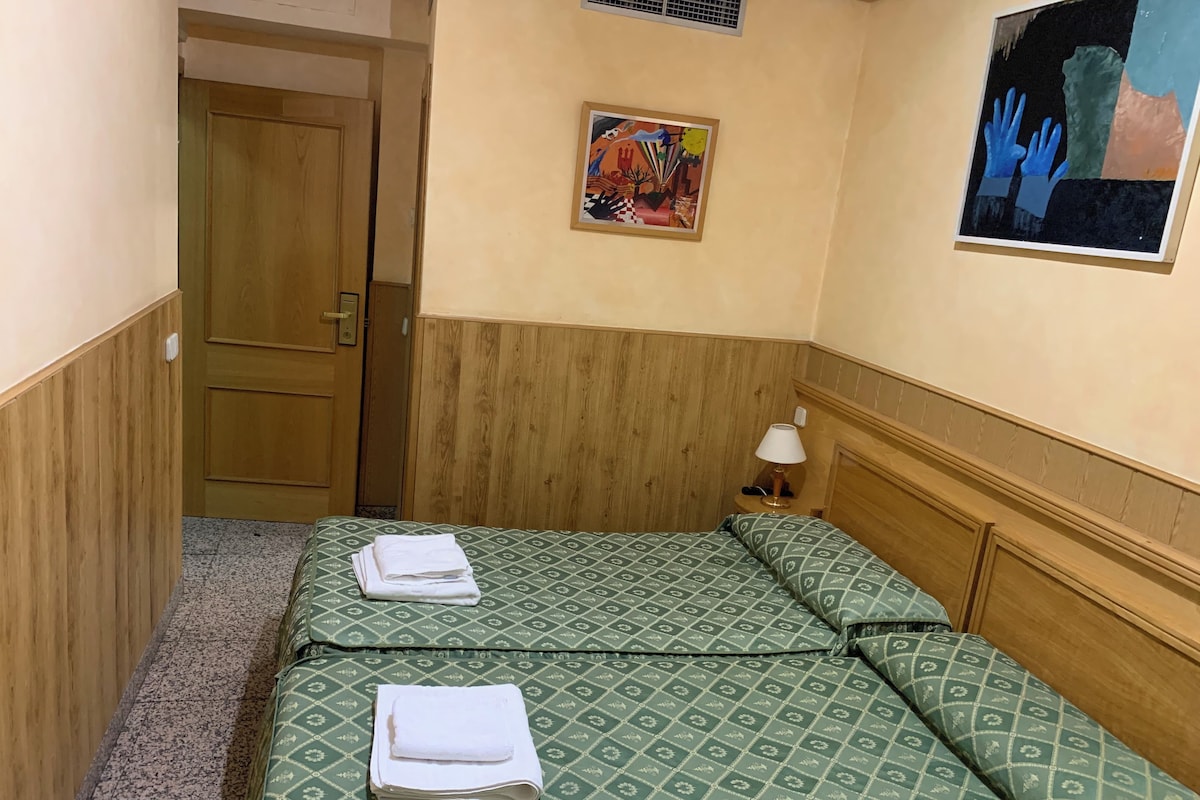 A hotel room with a bed, a nightstand, a lamp, a door, and a painting on the wall.