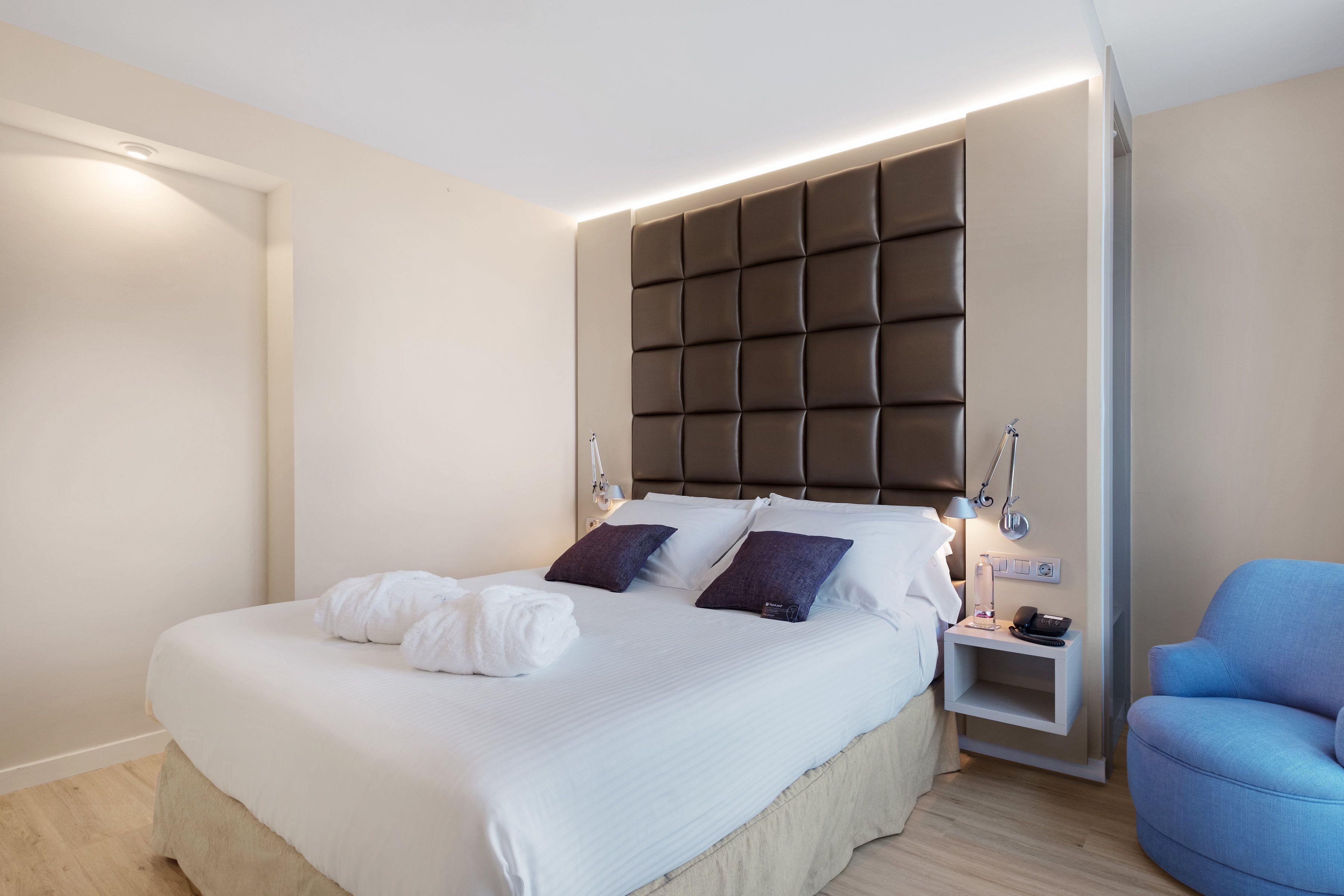 Una cama bien tendida con sábanas blancas y dos almohadas oscuras, una mesita de noche con una lámpara, y un sillón azul.