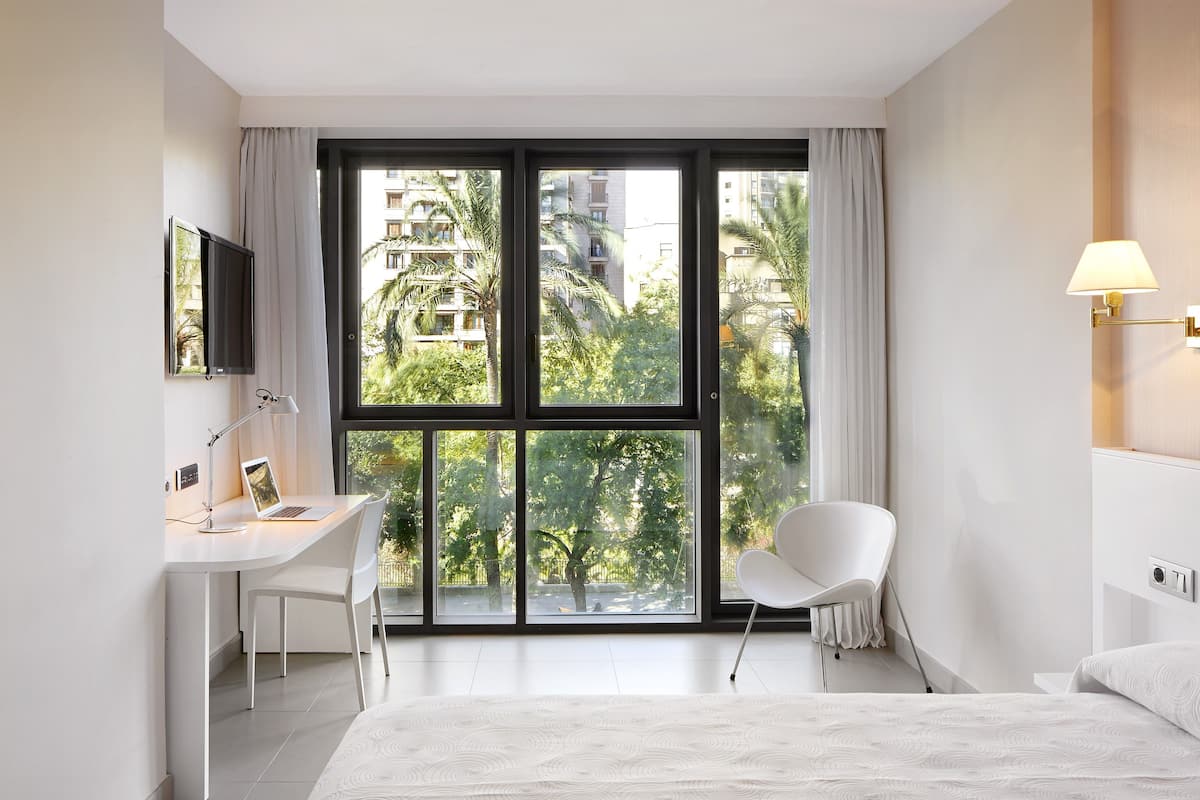 A modern hotel room with a large window, a desk with a chair, a bed, and a wall-mounted lamp.