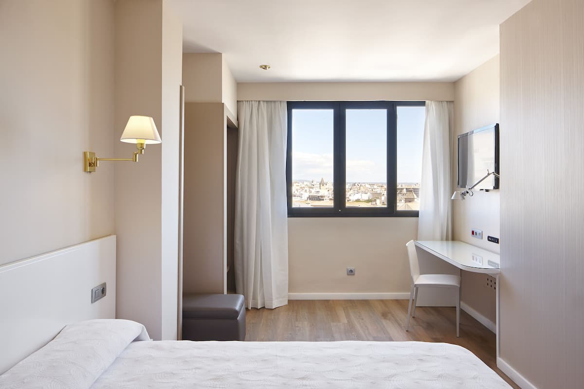 A hotel room with a bed, a desk, a chair, a TV, and a window with a city view.