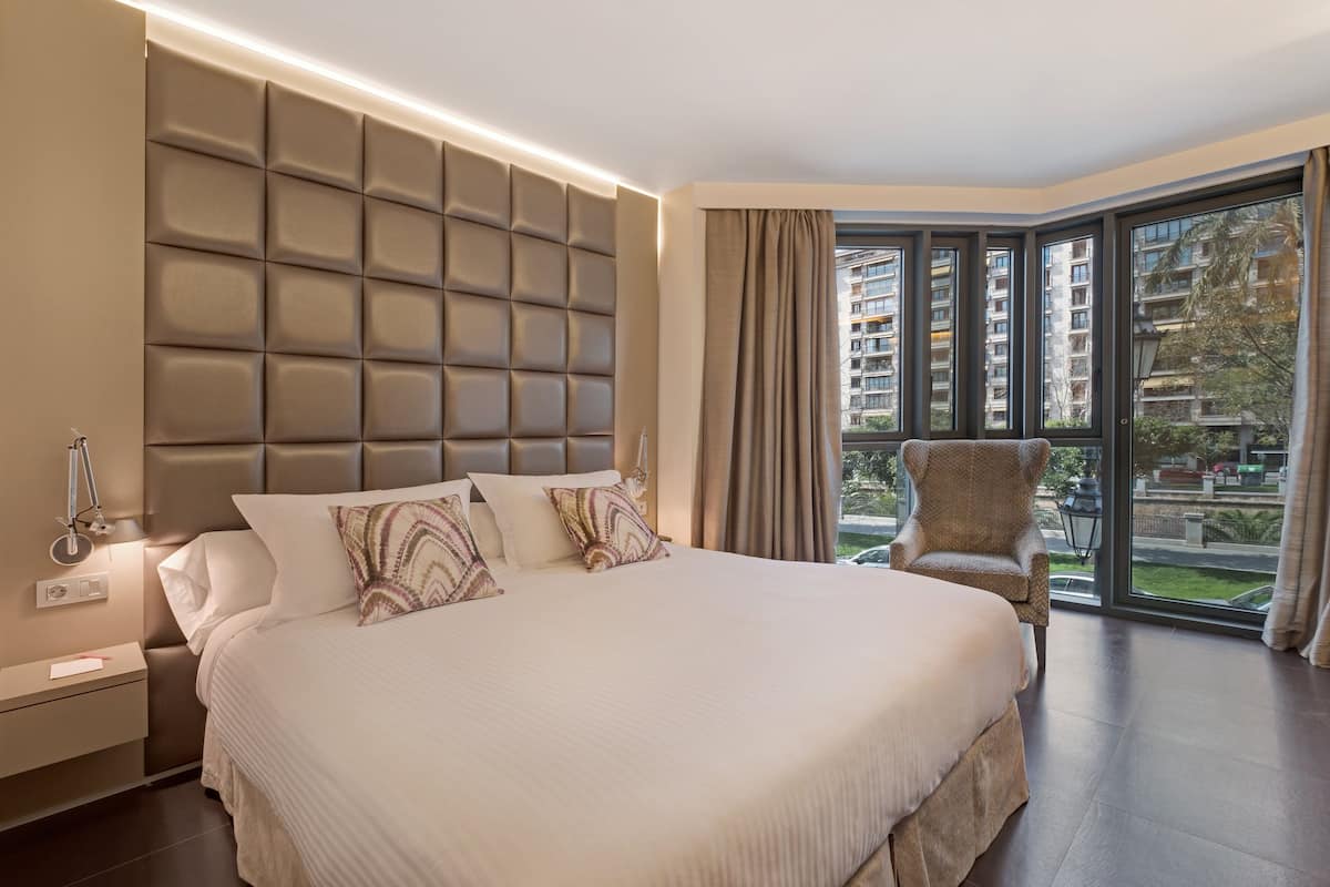 A modern hotel room with a large bed, a padded headboard, a chair, and a view of the city through large windows.