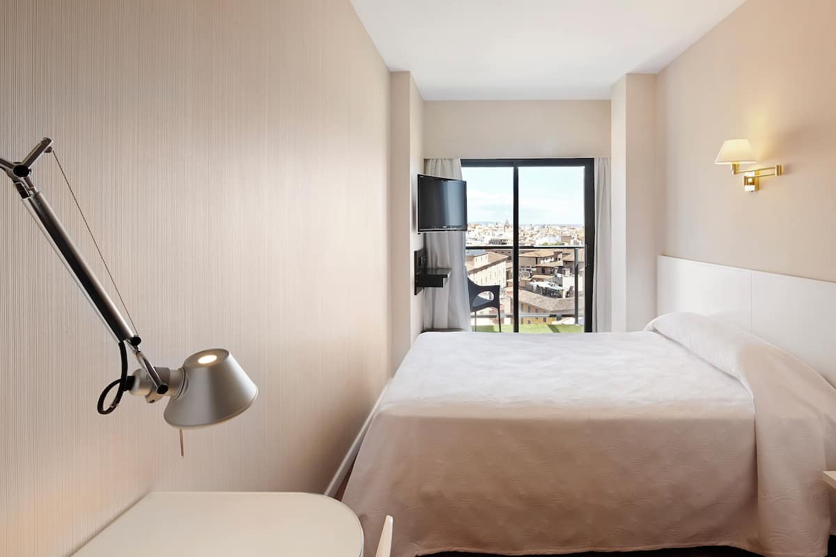 A hotel room with a bed, a desk, a chair, a TV, and a view of the city.