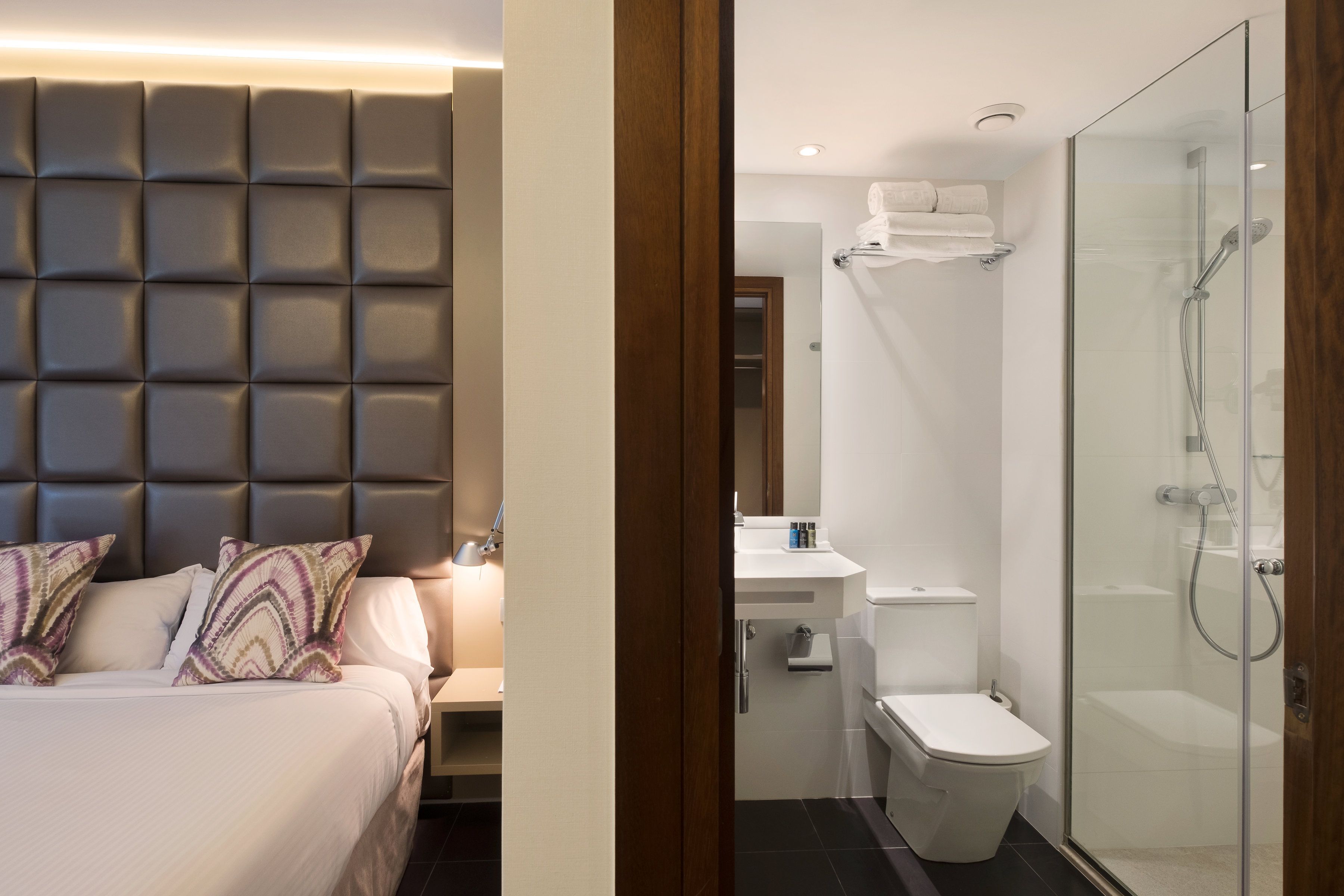 Une chambre à coucher moderne avec une tête de lit capitonnée, une table de chevet et une commode. À côté de la chambre se trouve une salle de bain dotée d’une douche à parois de verre, de toilettes et d’un lavabo.