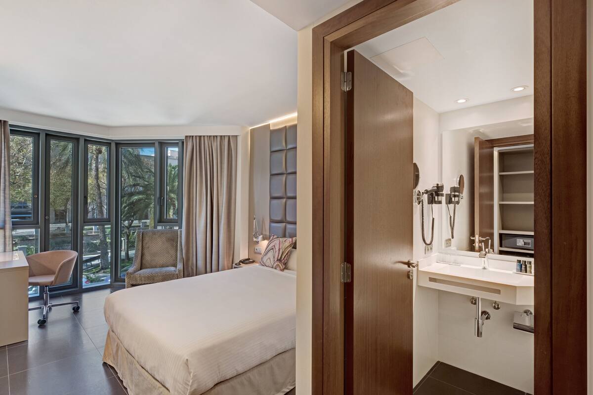 A modern hotel room with a large bed, a desk, a chair, and a bathroom with a vanity and mirror.