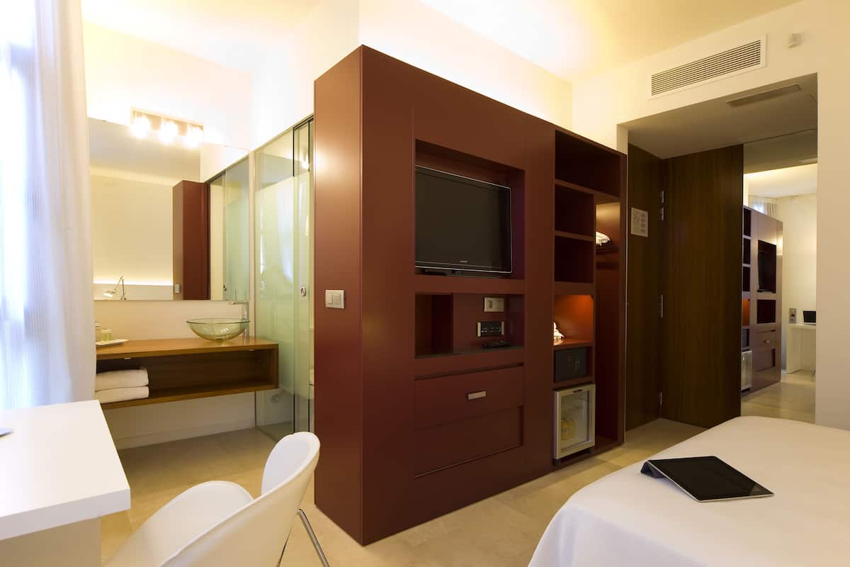 A modern hotel room with a large bed, a television, a glass-fronted bathroom, and a small desk with a chair.
