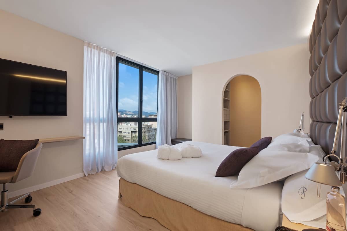 A modern hotel room with a large bed, a flat-screen TV, a desk, and a view of the city through large windows.