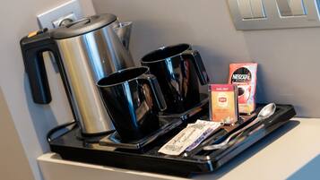 Een koffiehoek met een koffiezetapparaat, twee zwarte kopjes, een doos Nescafe, een pakje Lipton thee en een lepel op een dienblad.