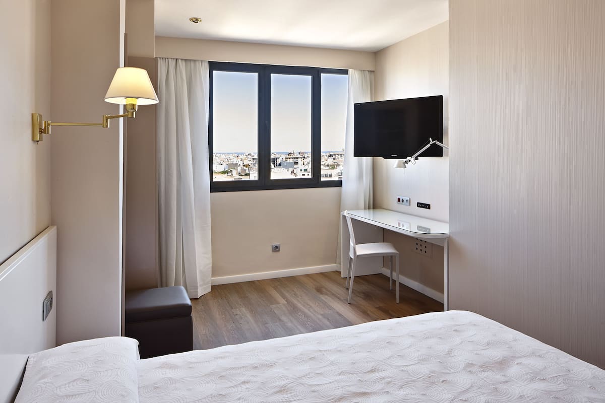 A hotel room with a bed, a desk, a chair, a TV, and a window with a city view.