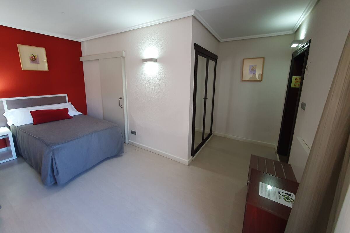 Un dormitorio con cama, mesita de noche, una puerta y una pequeña mesa con un cartel.