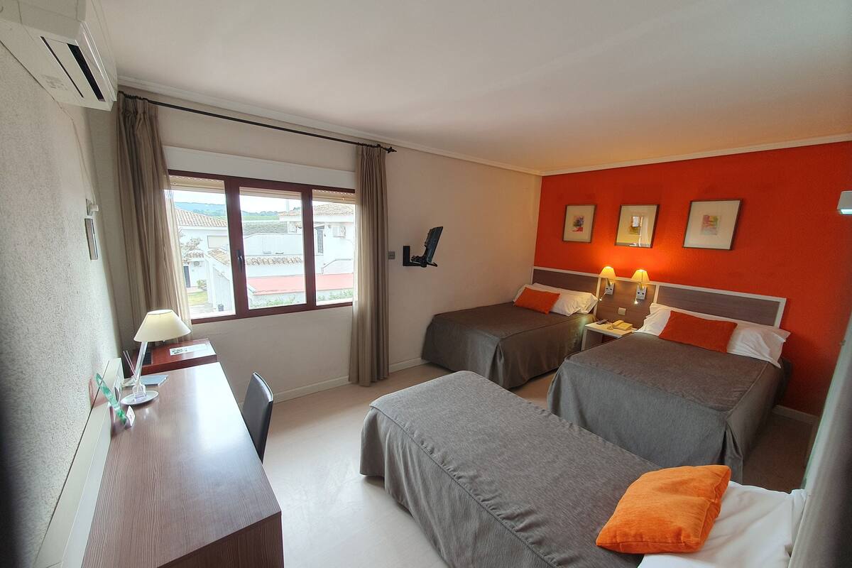 Habitación de hotel con dos camas individuales, un escritorio con silla, ventana con cortinas y vistas al exterior.