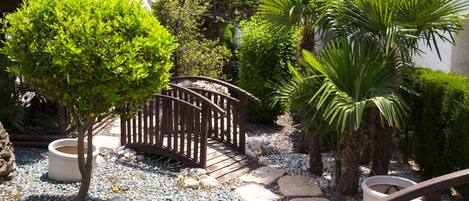 Un jardín con un sendero de piedra, un puente de madera y diversas plantas en macetas.