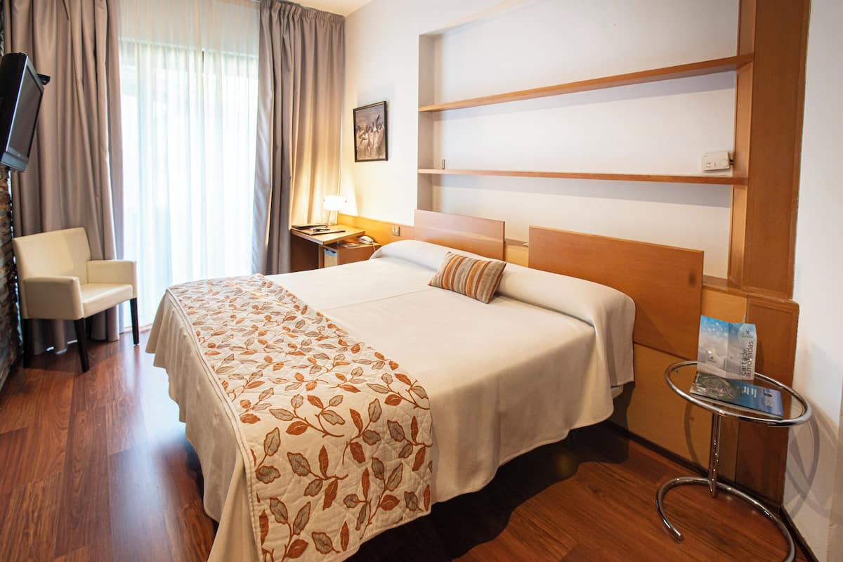 Quarto de hotel com cama, poltrona, escrivaninha com luminária, televisão e prateleira com um livro.