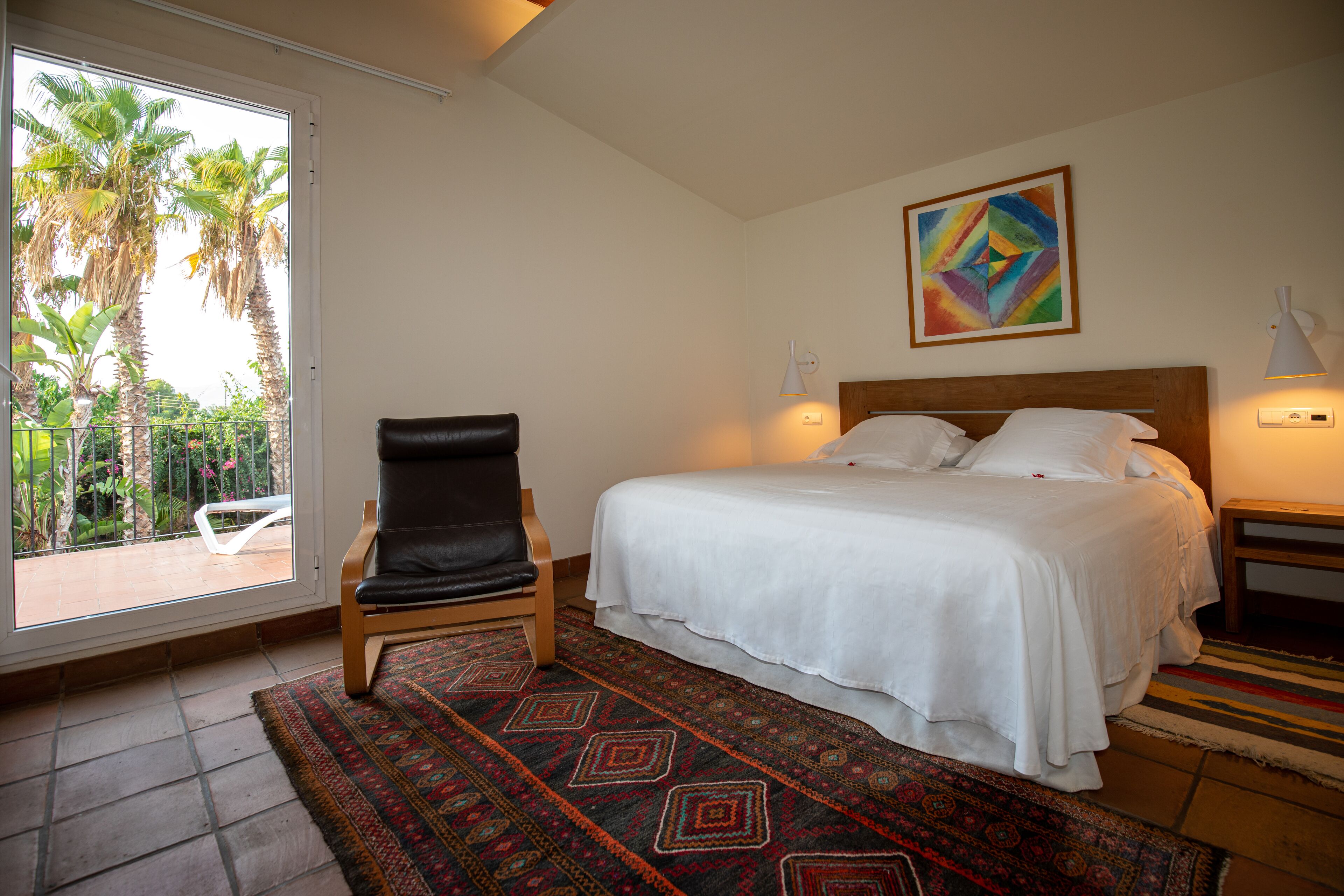 A hotel room with a bed, a chair, a colorful painting, and a view of palm trees through a glass door.